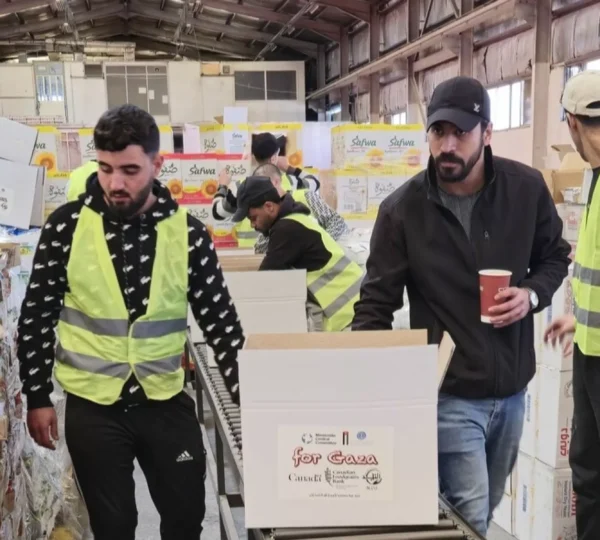 Several people in reflective vests organize and pack boxes in a warehouse filled with stacked goods. The scene conveys teamwork and purpose.