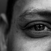 Close-up black and white photo of a person's eye, showing detailed textures of skin and eyebrow. The expression in the eye conveys introspection.