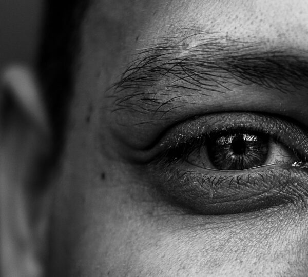 Close-up black and white photo of a person's eye, showing detailed textures of skin and eyebrow. The expression in the eye conveys introspection.