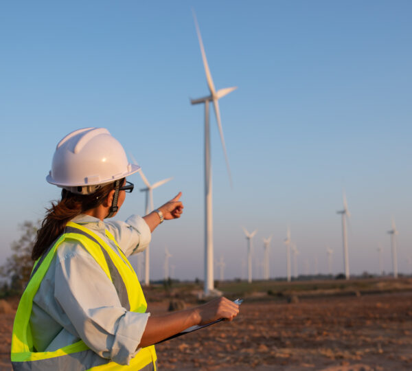 Female engineer in a hard hat and high-visibility vest pointing at wind turbines in a renewable energy farm during sunset.