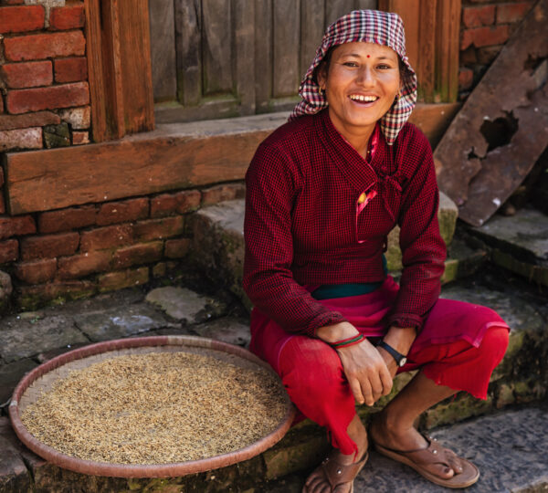 A person in a red traditional outfit and checkered headscarf smiles, sitting by a brick wall with a bamboo tray of grains. The scene conveys warmth and cultural richness.