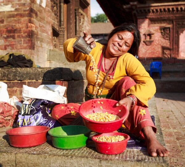 Smiling woman in traditional clothing sitting on stone steps beside a tray of harvested grains, in a rural village setting.