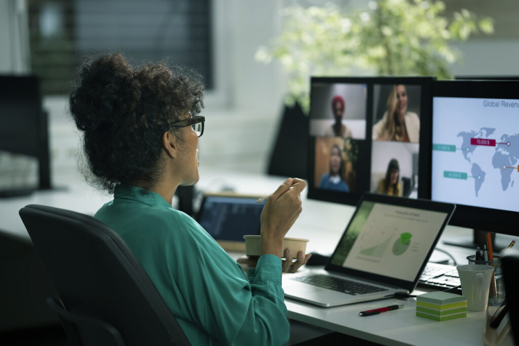 Woman in a green blouse participates in a video call at a desk, eating from a bowl. Multiple screens display charts and four people on a call.